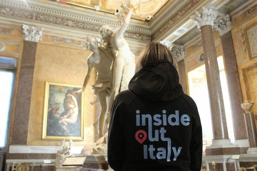 A person wearing a jacket with 'inside out Italy' text, standing in front of a classical statue in an ornate room with columns and a painting. 4