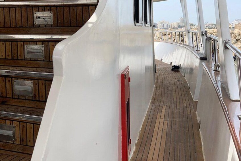 Ein Schiffdeck mit einem hölzernen Steg, Metallgeländern und einer Stadtansicht im Hintergrund. 2