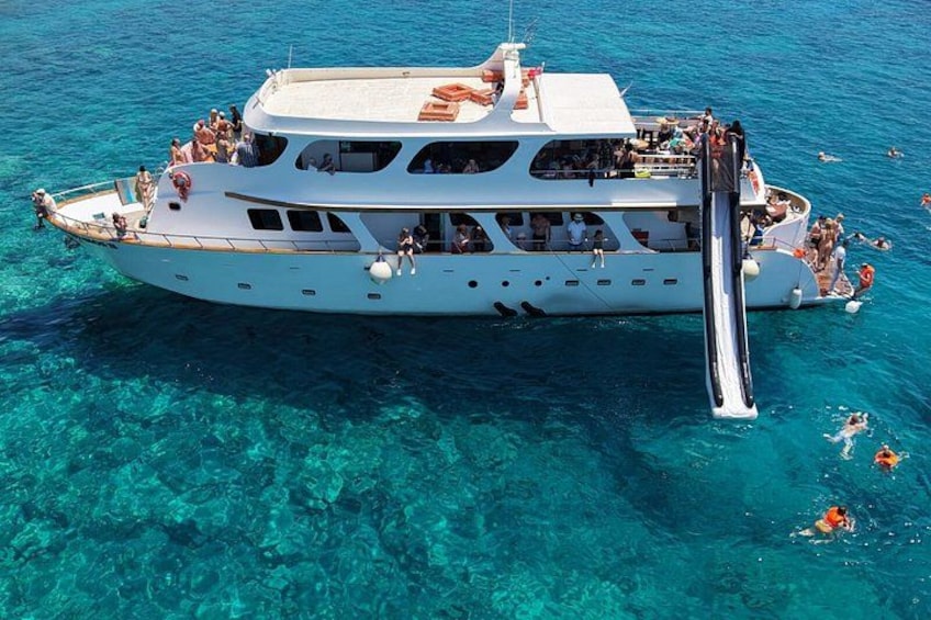 A large white boat with a slide, surrounded by clear blue water and people swimming. 2