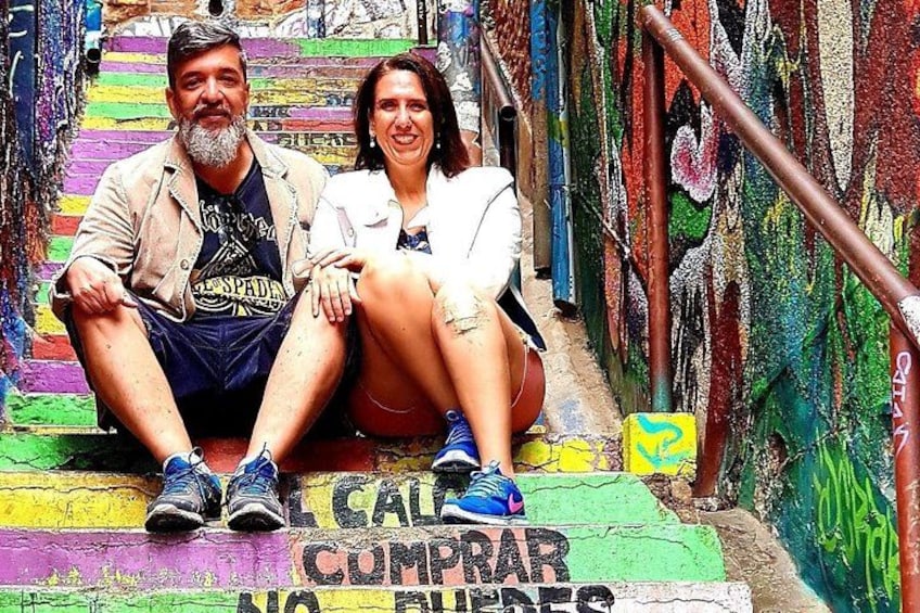 A couple sitting on a colorful staircase with graffiti and a sign that says 'CALLE COMPRAR NO FUELES'. 3