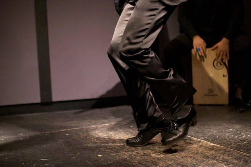 A person wearing black dress shoes with a shiny surface, standing on a dark floor with a visible stain. 2