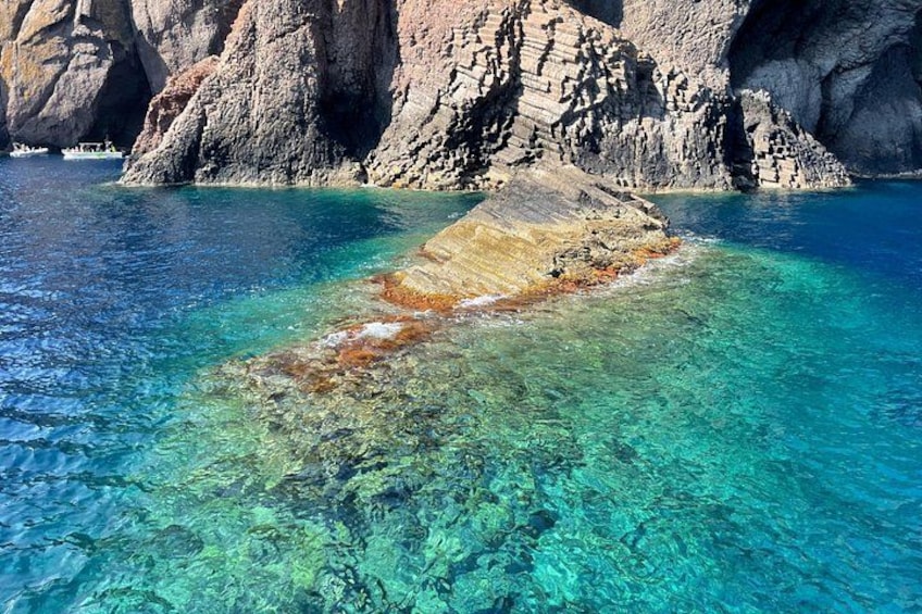 A clear blue sea with a rocky outcrop in the foreground and a larger rocky formation in the background. 2