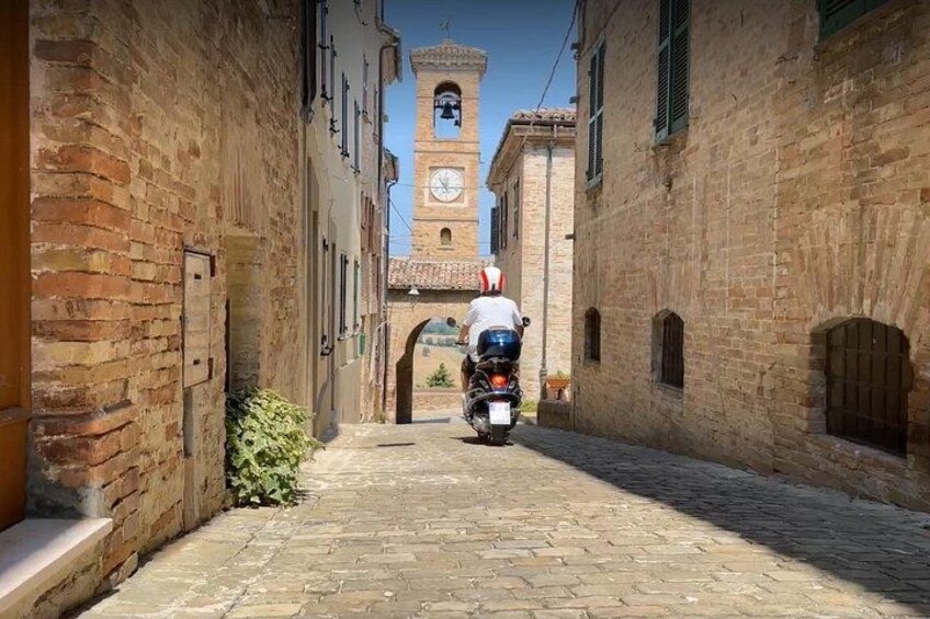 A narrow cobblestone street with a motorcyclist in the distance, flanked by brick buildings and a clock tower. 3