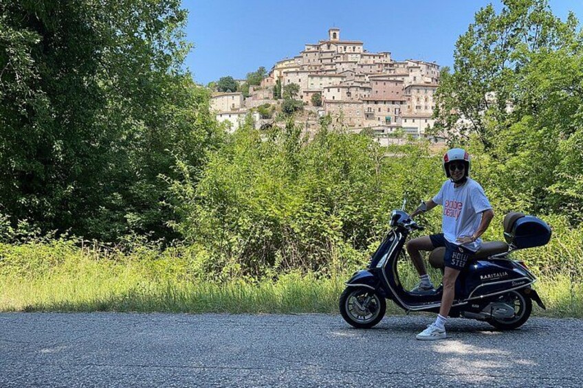 A person on a scooter, a road, and a hill with buildings in the background. 1