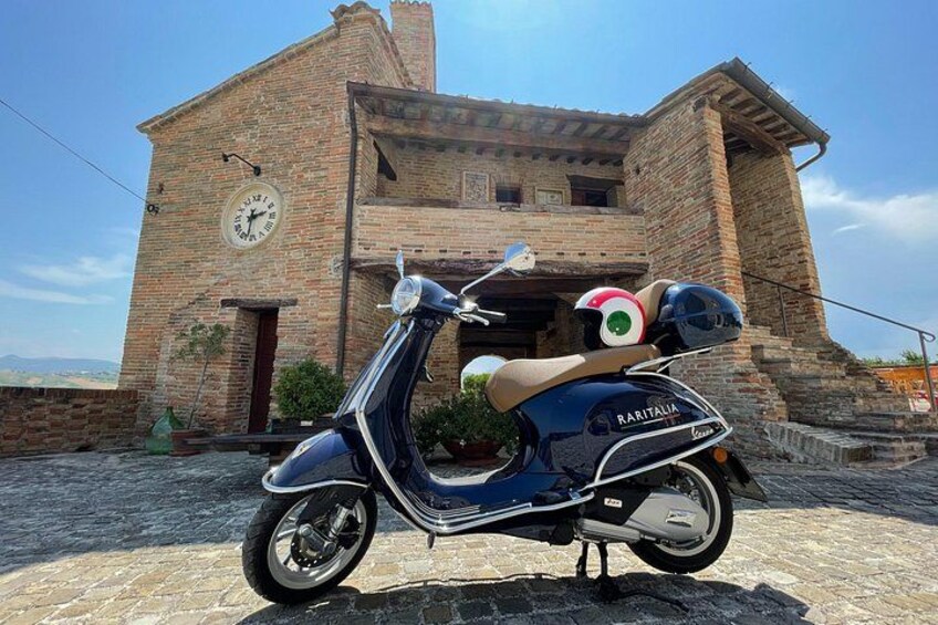 Ein restaurierter Vespa-Roller vor einem rustikalen Backsteinhaus mit einer Uhr. 2