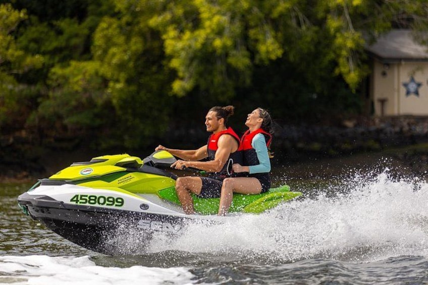 Two people on a jet ski, wearing life jackets, with a house and trees in the background. 3