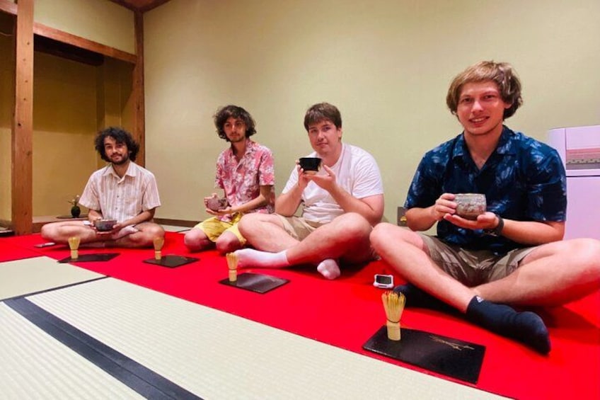 Four people sitting on tatami mats in a room with a red floor, holding cups. 3