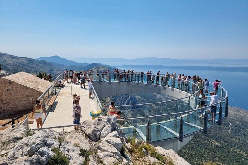 A glass-bottomed viewing platform with people overlooking a mountainous landscape and a body of water. 5