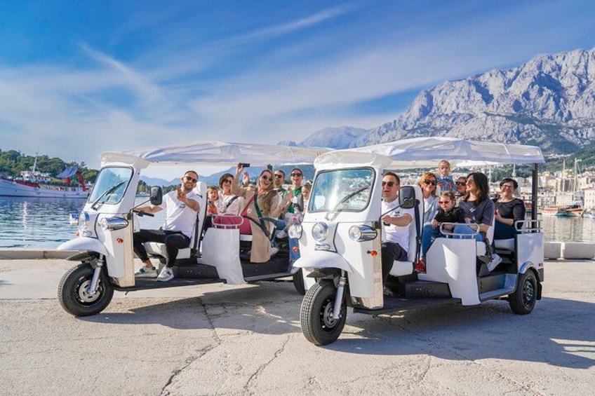 Two white three-wheeled vehicles with passengers, parked by a waterfront with mountains in the background. 2