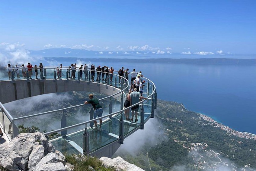 A glass observation deck with people observing a lake and distant mountains. 5