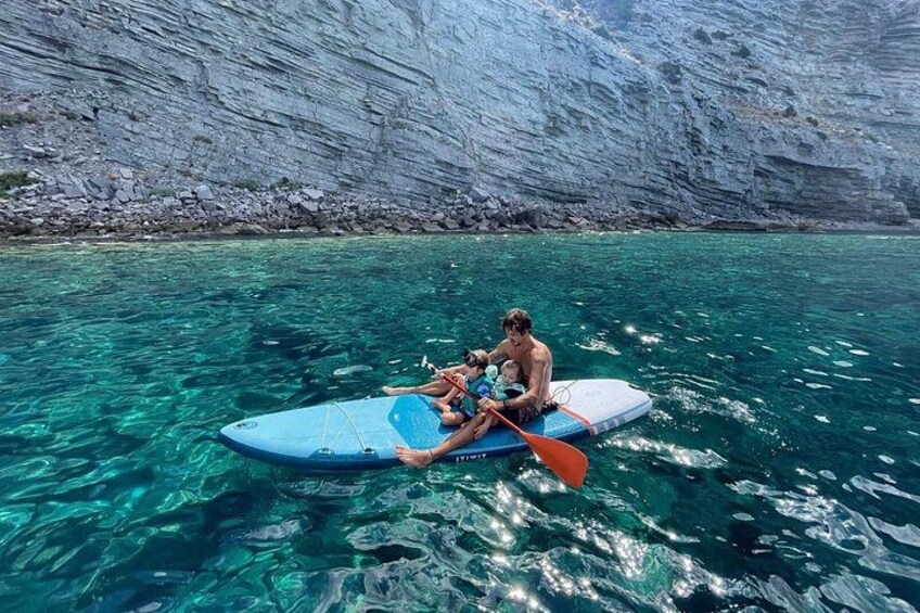 Een groep mensen op een blauw paddleboard in helder turquoise water, met een rotsachtige klif op de achtergrond. 4