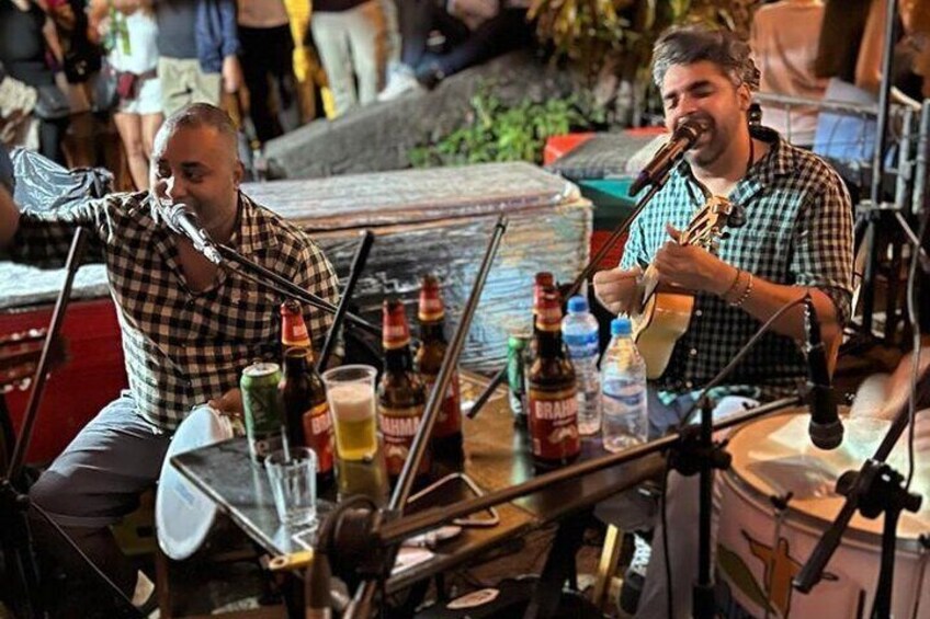 Two musicians performing at an outdoor event with musical instruments and bottles on the table. 1
