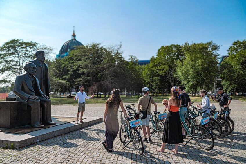 Een groep mensen, sommigen op fietsen, verzameld rond een standbeeld in een park, met een koepelvormig gebouw op de achtergrond. 4