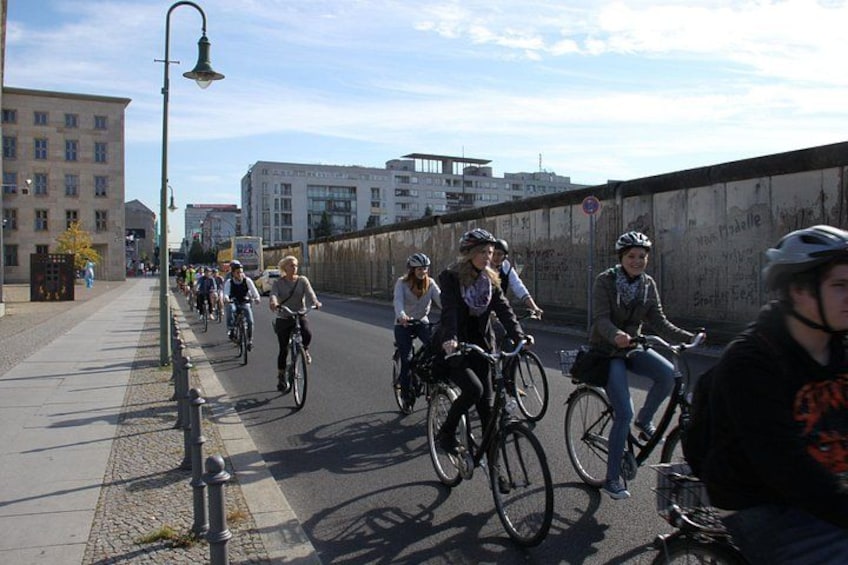 En grupp cyklister på en asfalterad cykelväg med byggnader och en mur i bakgrunden. 2