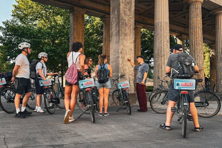 People standing around bicycles with signs, under a structure with columns. 4