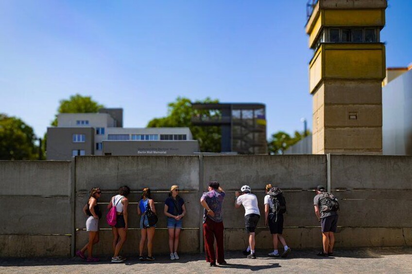 A group of people standing against a wall, some with backpacks, in an urban setting with buildings and a clear sky in the background. 5