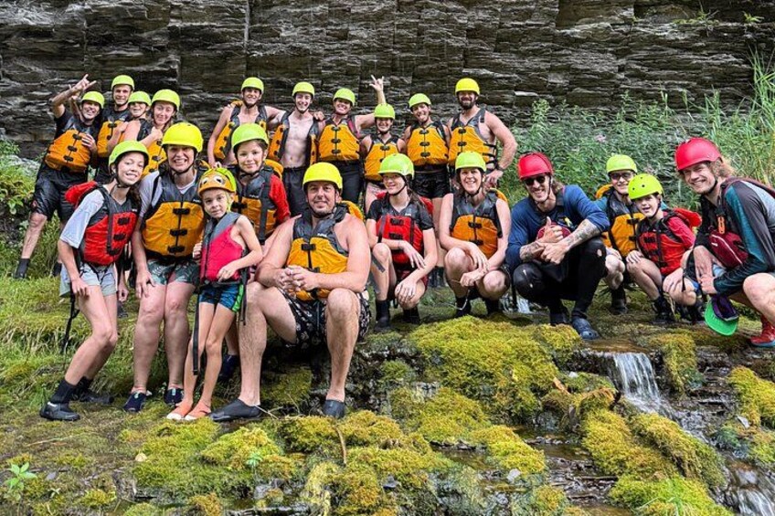 A group of people wearing life jackets and helmets, posing in a natural setting with moss-covered rocks and a small stream. 4