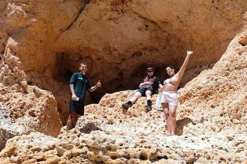 Three people in a rocky outdoor setting with a large cave-like structure in the background. 3