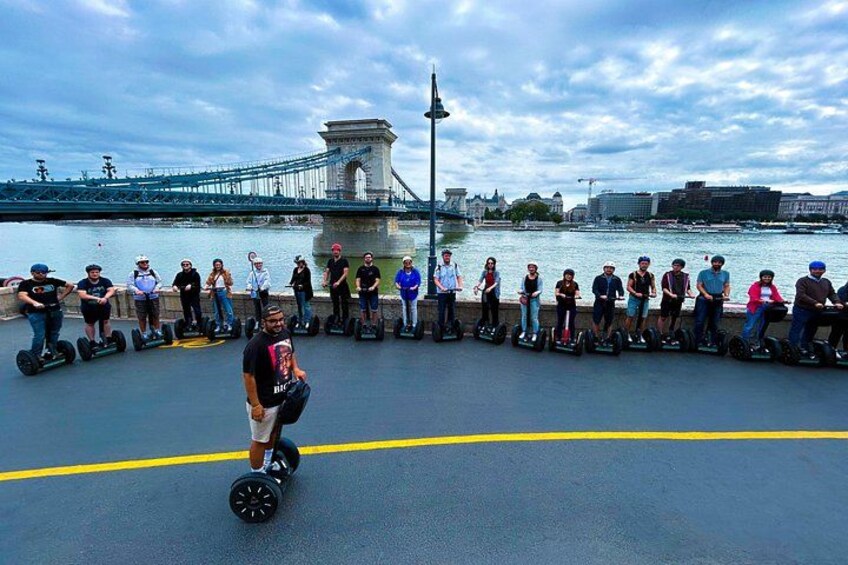 Un groupe de personnes sur des gyropodes, au bord d’une rivière, avec un pont en arrière-plan. 3