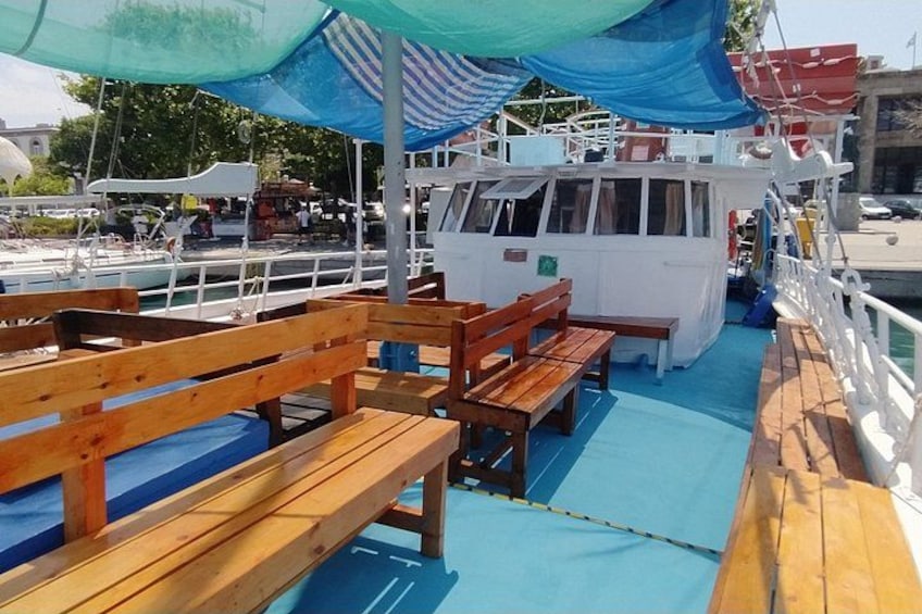 A boat with wooden benches and a blue floor, docked at a marina with other boats and a building in the background. 3