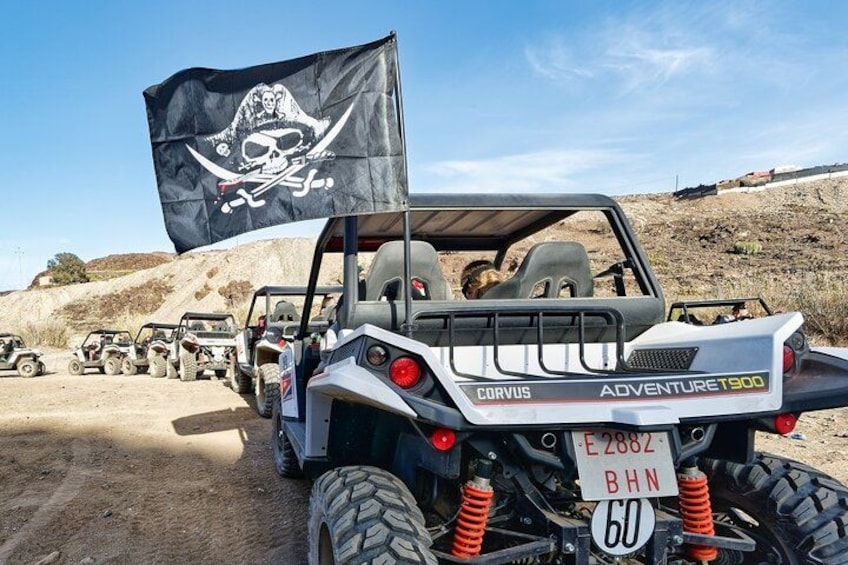 A white off-road vehicle with a pirate flag, labeled 'CORVUS ADVENTURE T900,' parked on a dirt road with other similar vehicles in the background. 1