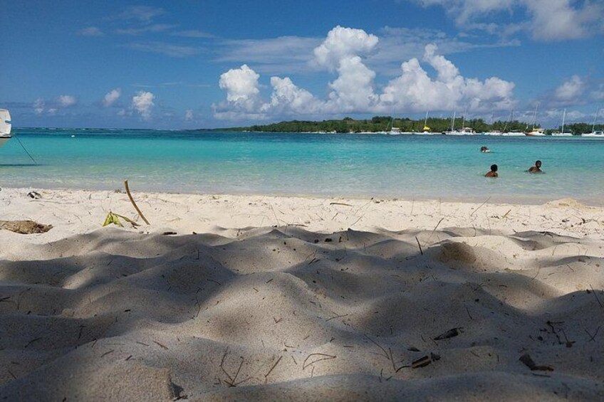 A sandy beach with clear turquoise water, a few people swimming, and boats in the distance. 5
