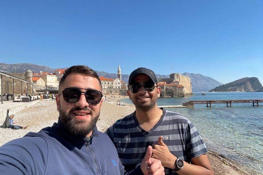 Due persone si fanno un selfie su una spiaggia, con vista chiara su un edificio storico e montagne sullo sfondo. 3