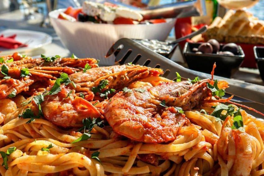 Un plato de pasta con mariscos, camarones y pasta, decorado con hierbas, servido sobre una mesa con un bol de aceitunas y pan al fondo. 2