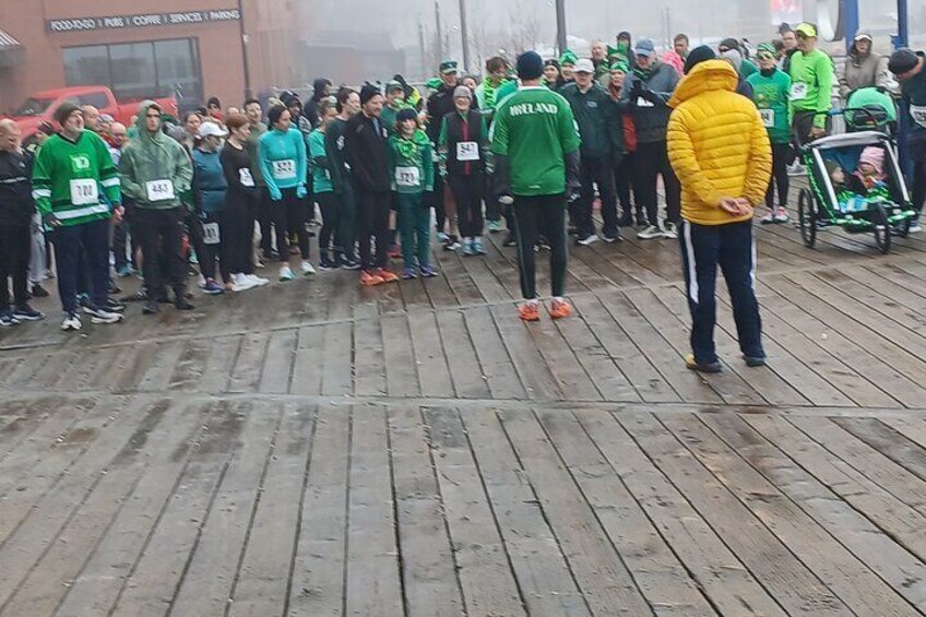 A group of people standing on a wooden deck, some wearing numbered bibs, likely at a race or event. 5