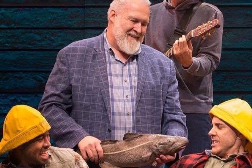 Ein Mann in einem karierten Blazer hält einen großen Fisch, im Vordergrund sind zwei Personen mit gelben Hüten zu sehen. 5