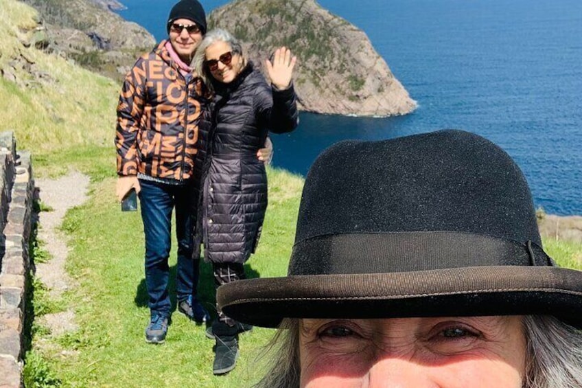En person med svart hatt och kappa som står på en gräsbevuxen stig, med en klippig brant och havet i bakgrunden. 2