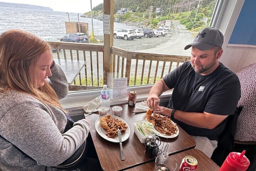Dos personas cenando en una mesa con comida y bebidas, con vista a un estacionamiento y una masa de agua. 1