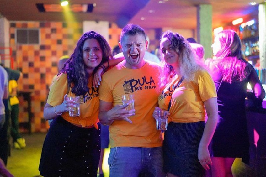 People wearing matching orange shirts with text, holding drinks, and a bar in the background. 5