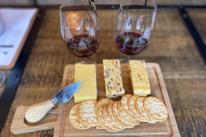 A wooden cutting board with cheese, crackers, and a knife. Two glasses of red wine are in the background. 5