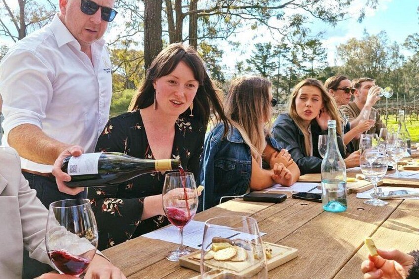 A group of people at an outdoor picnic with a wooden table, wine glasses, and a bottle being poured. 1