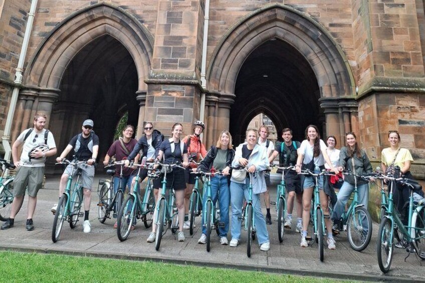 A group of people with bicycles in front of a historic stone building with arches. 3