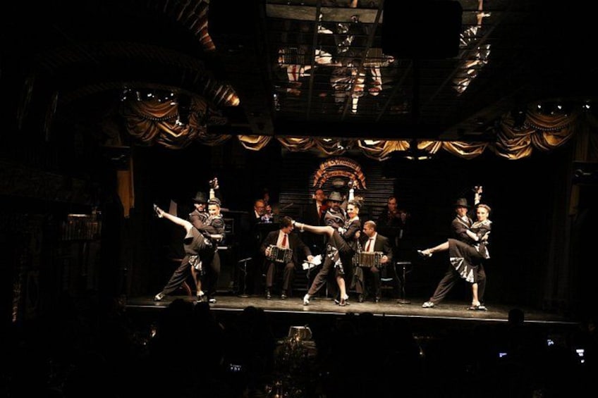 A stage with performers in period costumes, a chandelier, and a backdrop with a decorative arch. 1