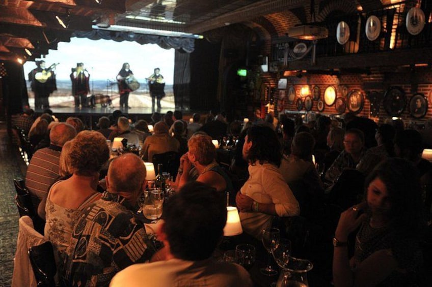 A crowded bar with people seated at tables, a stage with performers, and a brick wall with clocks. 2