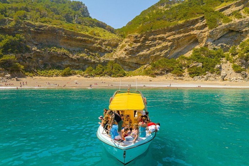 Un bateau surmonté d’un auvent jaune, naviguant sur une eau turquoise et limpide, entouré de falaises rocheuses et d’une plage de sable. 2