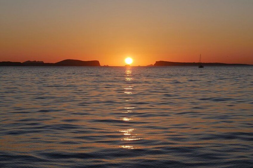 A sunset over a calm sea with a sailboat and distant landforms. 2