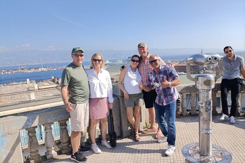 Een groep mensen staat nabij een historische uitkijktoren met uitzicht op de haven en verre bergen. 3