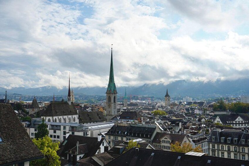 Eine Stadtansicht mit historischen Gebäuden, einem markanten Kirchturm und Bergen im Hintergrund. 1