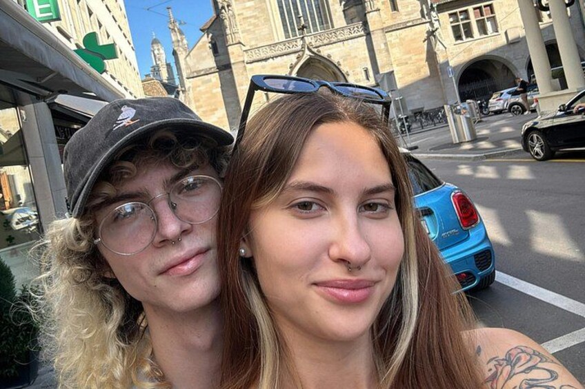 Zwei Personen machen ein Selfie auf einer Stadtstraße, im Hintergrund ein blaues Auto und historische Gebäude. 4