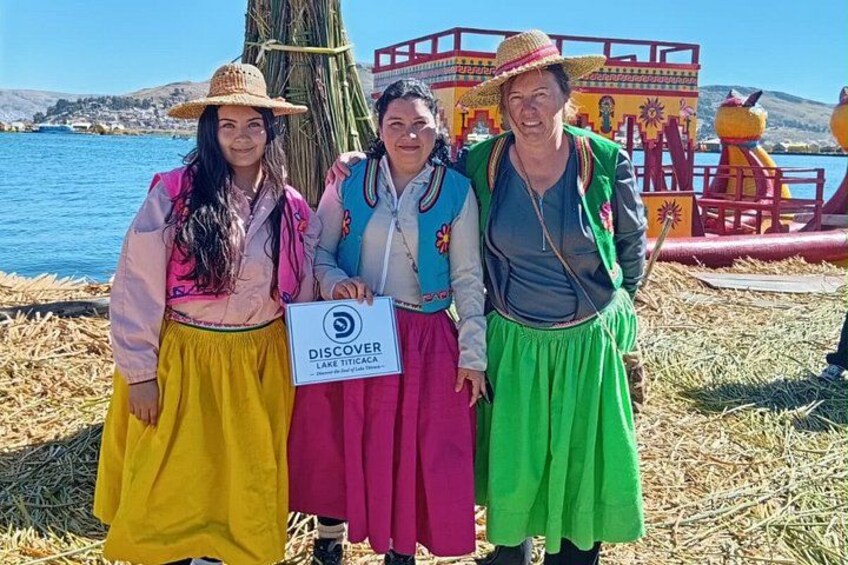 Drie mensen in traditionele kledij staan voor een kleurrijk gebouw met een bord waarop staat 'DISCOVER LAKE TITICACA'. 1