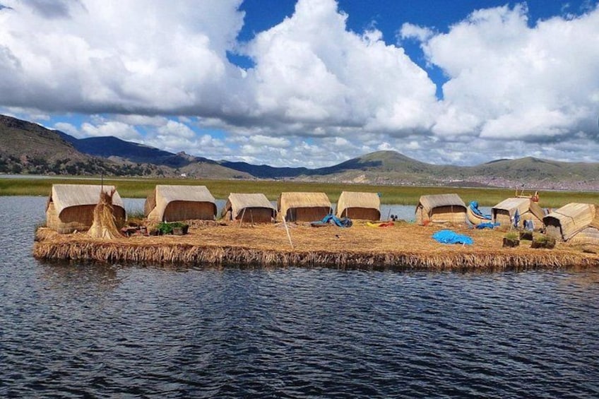 A series of thatched huts on a floating platform in a body of water, surrounded by hills and a partly cloudy sky. 2