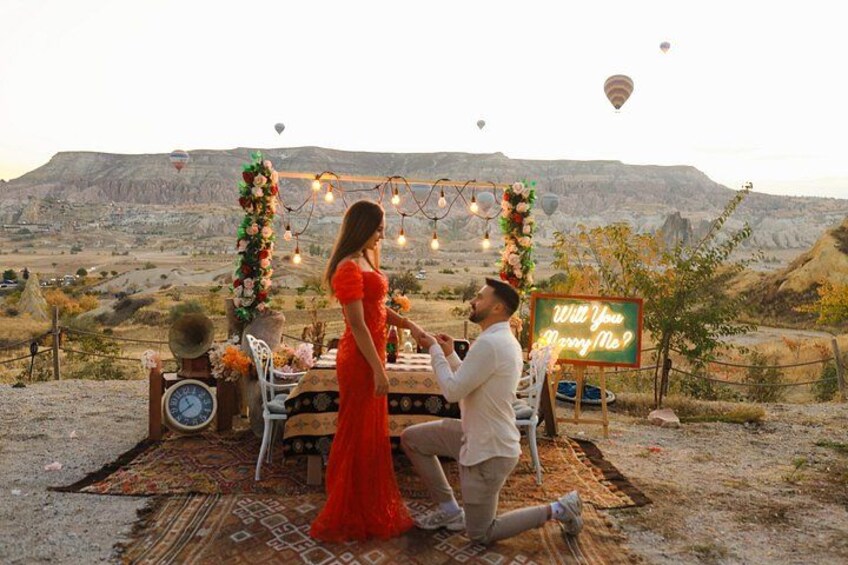 A couple at a table with a sign reading 'Will You Marry Me?' in a desert landscape with mountains in the background. 2