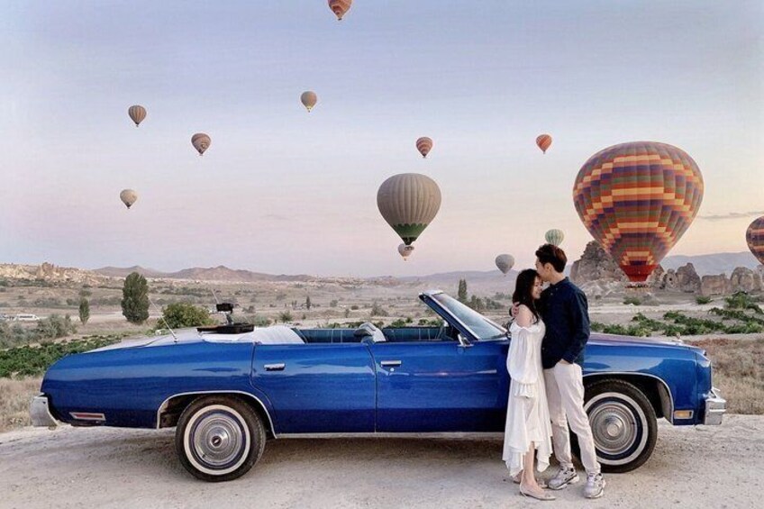 A blue convertible car parked on a dirt road with a couple standing beside it. Hot air balloons are visible in the background. 2