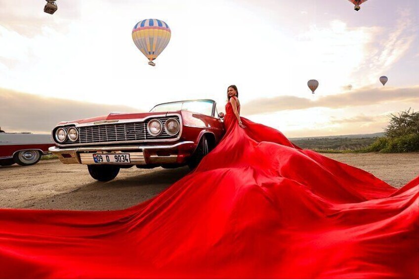 A vintage red car with a woman in a red dress standing next to it. Hot air balloons are visible in the background. 1