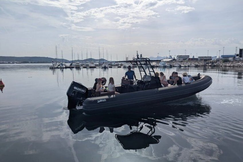 A boat with passengers on a calm body of water, surrounded by other boats and a distant shoreline. 3