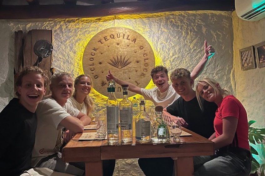 Un grupo de personas sentadas a una mesa con botellas y vasos, un reloj y una pared con un diseño circular. 3